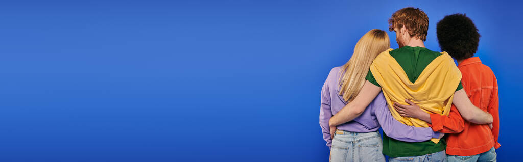 Liebesdreieck, Polyamory, drei Personen, Rückansicht eines rothaarigen Mannes, der multirassische Frauen auf blauem Hintergrund umarmt, Studioaufnahme, lebendige Farben, lässige Kleidung, stilvolle Kleidung, moderne Familie, Banner  - Foto, Bild