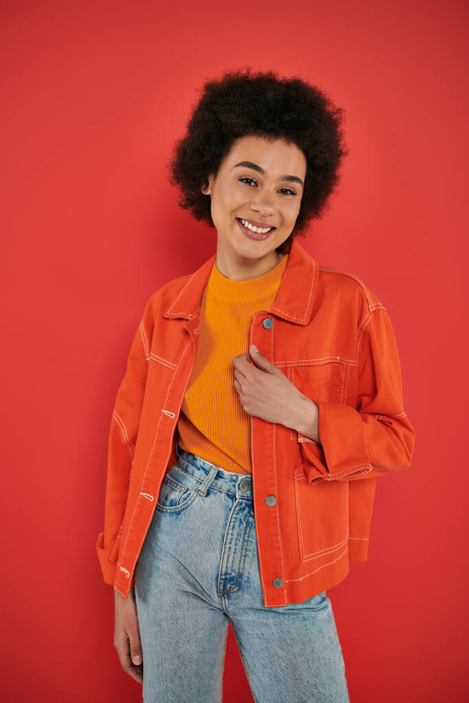happy young african american woman posing in stylish and vibrant outfit on coral background, smiling and looking at camera, fashion statement, African beauty, natural curly hair, trend  - Photo, Image