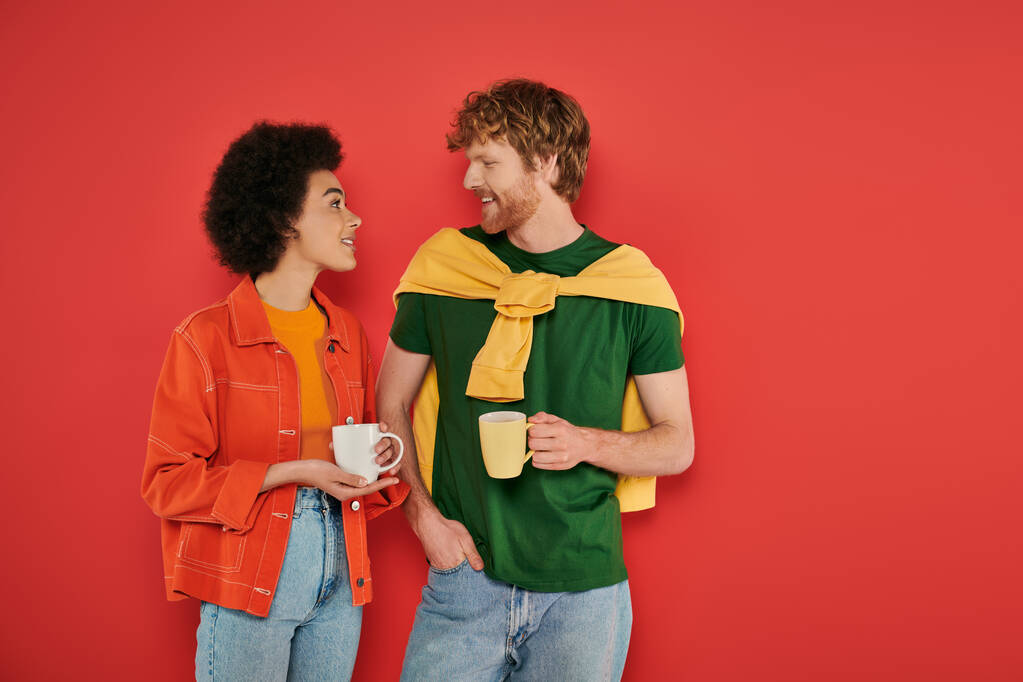 мультикультурна пара тримає чашки кави на кораловому фоні, ранкова рутина, культурне різноманіття, яскраві кольори, стильний одяг, міжрасові коханці тримають чашки і розмовляють  - Фото, зображення
