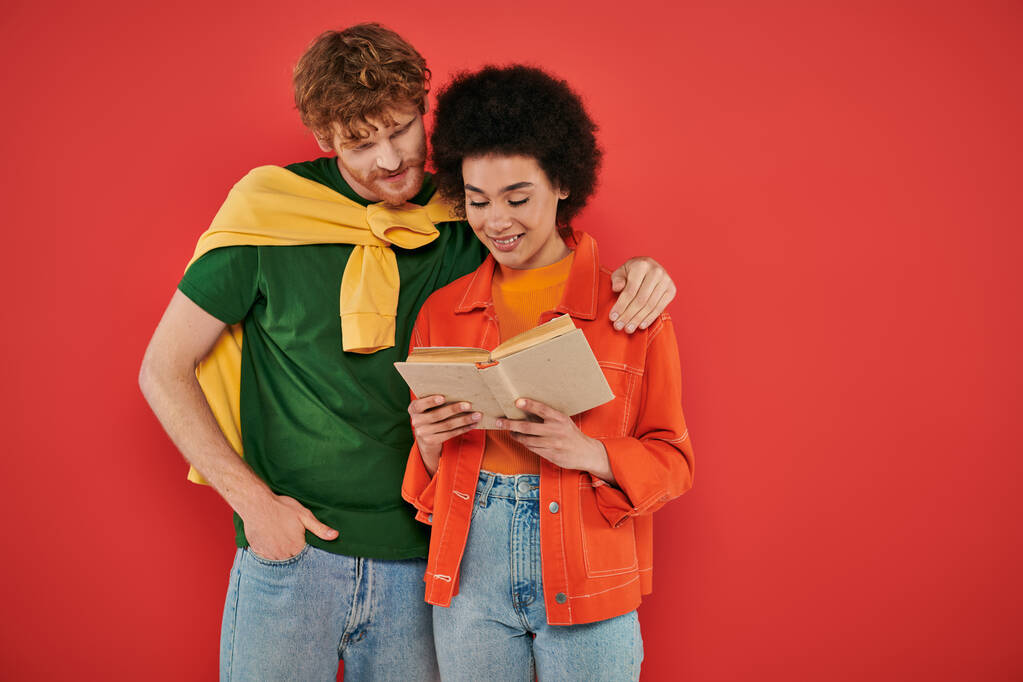 interracial couple hugging and reading book on coral background, cultural diversity, vibrant colors, stylish outfits, youth and intelligence, multicultural people spending lovely time together - Photo, Image