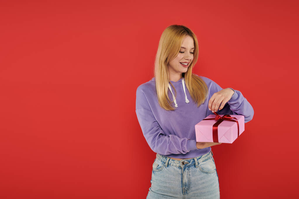 holiday, happiness, blonde young woman in casual attire opening present on coral background, vibrant colors, wrapped gift box, attractive and stylish, festive occasions, purple hoodie and jeans  - Photo, Image
