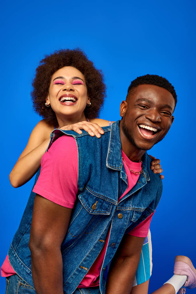 Wesoła młoda Afroamerykanka z odważnym makijażem przytulając najlepszą przyjaciółkę w stylowym stroju i bawiąc się stojąc razem na niebiesko, najlepsi przyjaciele w dopasowanych strojach, przyjaźń - Zdjęcie, obraz