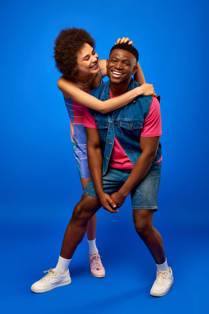 Полная длины позитивная молодая африканская женщина в раздевалке обнимает лучшую подругу в летнем стильном наряде, стоя и веселясь на голубом фоне, лучшие друзья хорошо проводят время - Фото, изображение