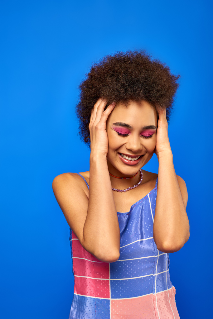 Portrait of smiling stylish young african american woman with natural hair and bold makeup touching head while standing and posing isolated on blue, charismatic model in summer outfit - Photo, Image