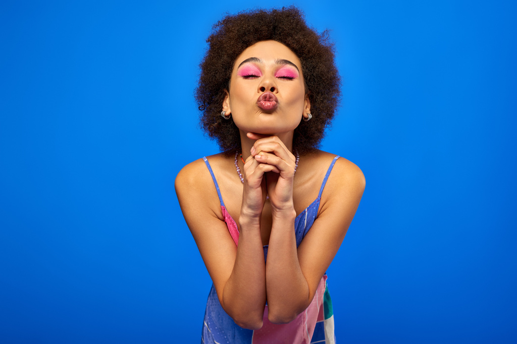 Retrato de jovem afro-americana com maquiagem elegante e ousada vestindo vestido de verão e lábios amuados enquanto posando e de pé isolado em azul, modelo carismático em roupa de verão - Foto, Imagem