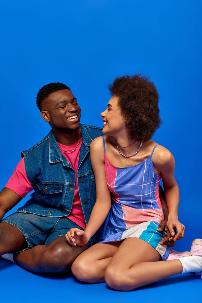 Νέοι και στιλάτοι αφροαμερικανοί καλύτεροι φίλοι με καλοκαιρινά ρούχα χαμογελώντας κοιταζόμενοι, περνώντας χρόνο μαζί και καθισμένοι σε μπλε φόντο, στιλάτοι φίλοι ποζάρουν με αυτοπεποίθηση - Φωτογραφία, εικόνα
