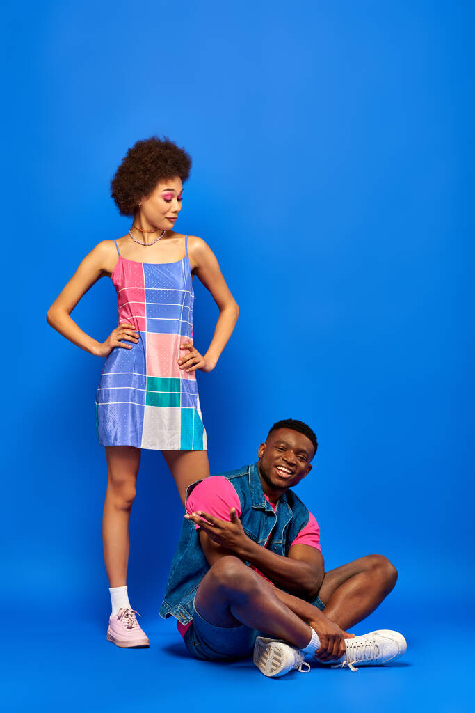 Full length of astonished young african american woman with bold makeup and sundress looking at stylish best friend smiling and sitting on blue background, stylish friends posing confidently - Photo, Image