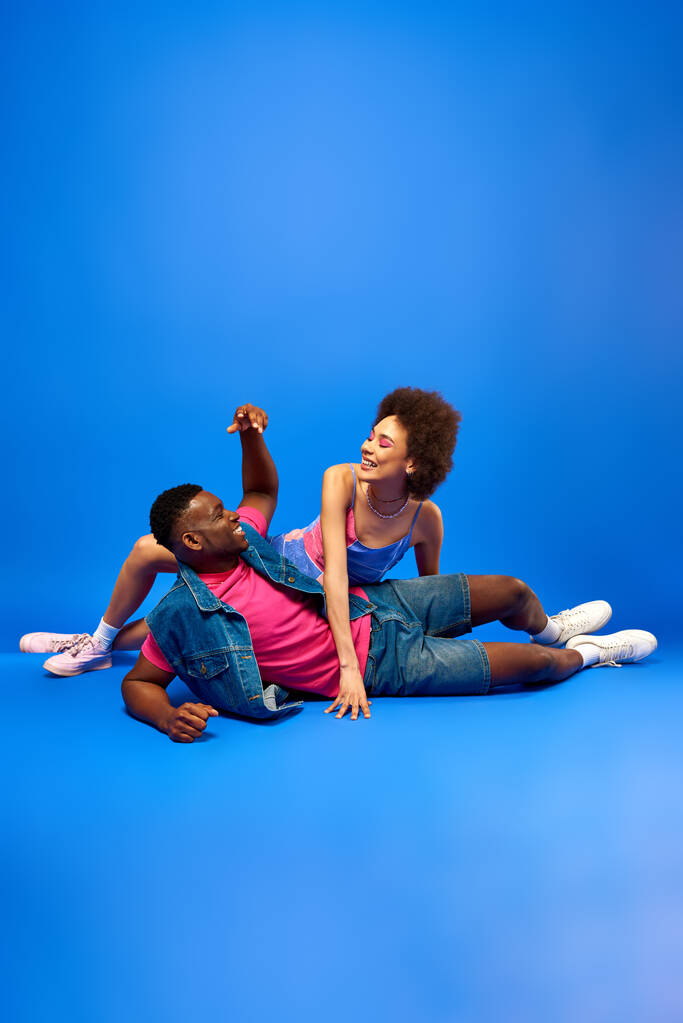 Pozitivní mladá africká americká žena s tučným make-upem v slunečních šatech pózuje poblíž stylové nejlepší kamarádky v džínové vestě a tričku na modrém pozadí, módní bestie vyzařující důvěru  - Fotografie, Obrázek