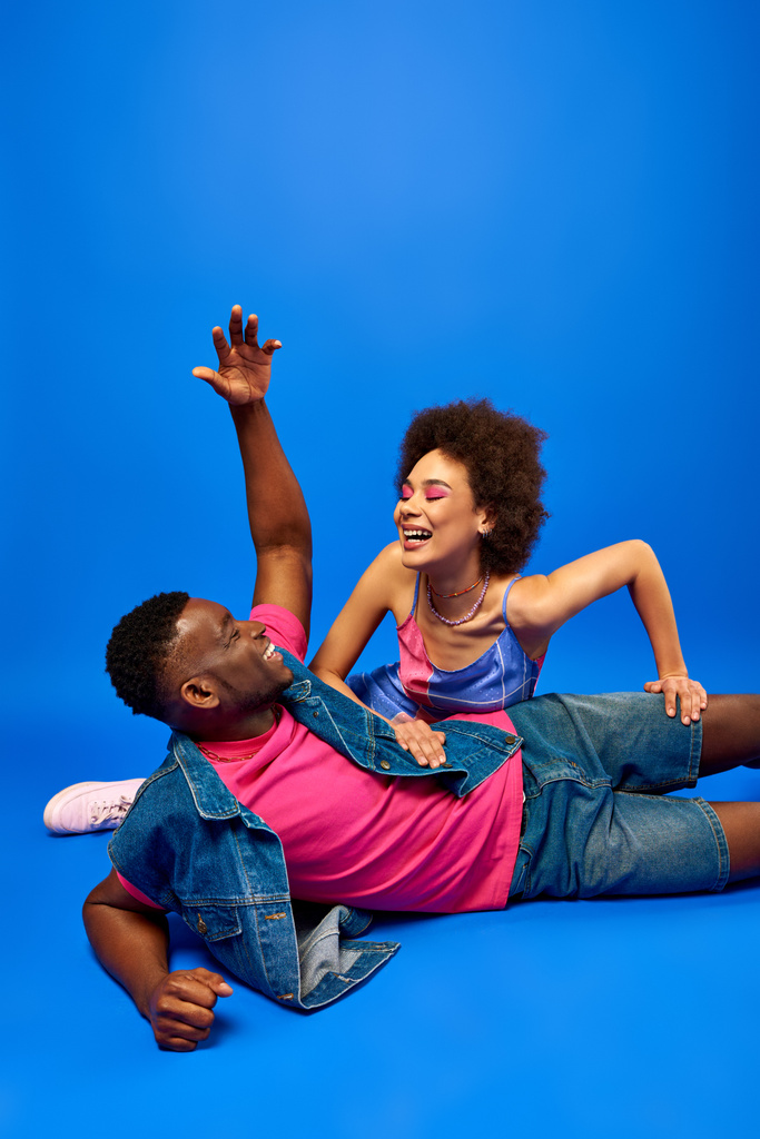 Веселый молодой африканский американец с современной прической разговаривает с лучшим другом с смелым макияжем и сарафаном, сидя на синем фоне, модные подруги излучают уверенность  - Фото, изображение