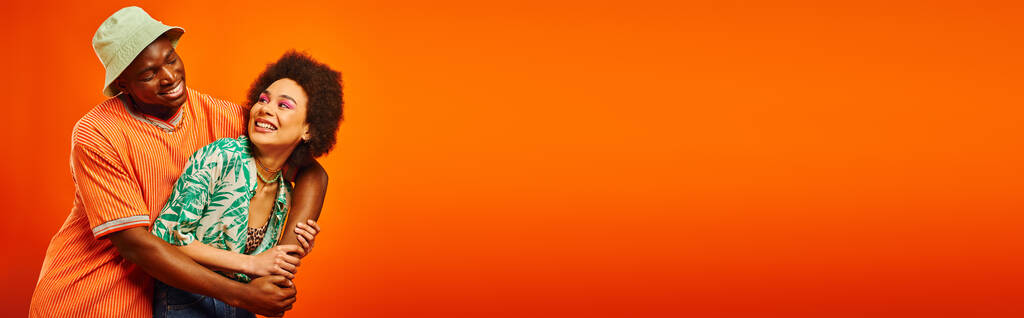 Allegro giovane uomo afroamericano in abito estivo e cappello panama che abbraccia il miglior amico con trucco audace e in piedi isolato su arancione, amici in mostra stile individuale, banner  - Foto, immagini