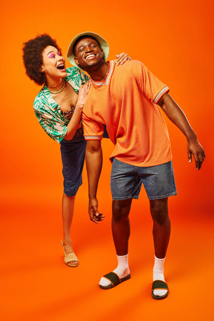 Comprimento total de excitado jovem afro-americana em roupa de verão abraçando alegre melhor amigo em chapéu panama, enquanto em pé sobre fundo laranja, amigos apresentando estilo individual - Foto, Imagem