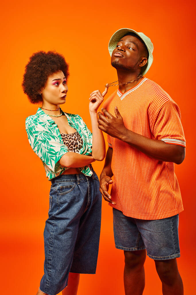 Скептически настроенная африканская американка со смелым макияжем, трогательной цепью на лучшего друга в панамской шляпе и летней одежде, изолированной от оранжевого, друзей, демонстрирующих индивидуальный стиль, концепцию дружбы - Фото, изображение