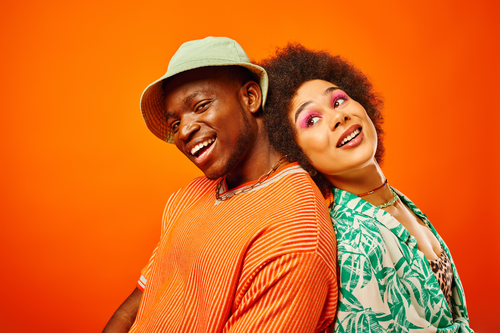 Vidám fiatal afro-amerikai férfi panama kalapban a kamerába néz, miközben háttal áll a legjobb barátjának nyári ruhában együtt elszigetelve a narancssárga, barátok divatos ruhákban - Fotó, kép