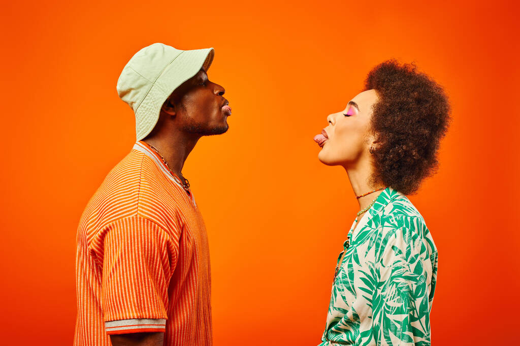 Side view of young and trendy african american best friends in summer outfits sticking out tongues while standing together isolated on orange, friends in trendy clothes, friendship concept - Photo, Image