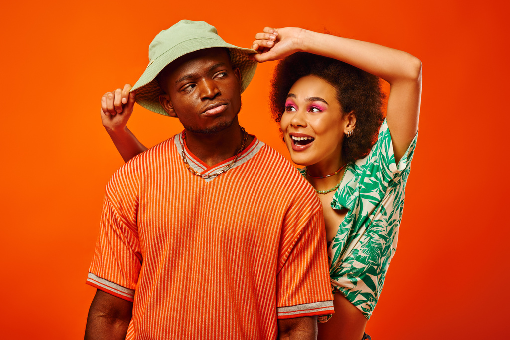 Usmívající se mladá afro-americká žena s tučným make-upem v letních šatech dotýkající se panamského klobouku vážného nejlepšího přítele a stojící osamoceně na oranžové, přátelé v módních šatech, přátelství - Fotografie, Obrázek