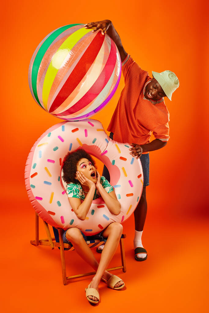 Positieve jonge Afrikaan Amerikaanse man in trendy zomer outfit houden bool bal in de buurt geschokt beste vriend met zwembad ring op dek stoel op oranje achtergrond, fashion-forward vrienden, vriendschap  - Foto, afbeelding