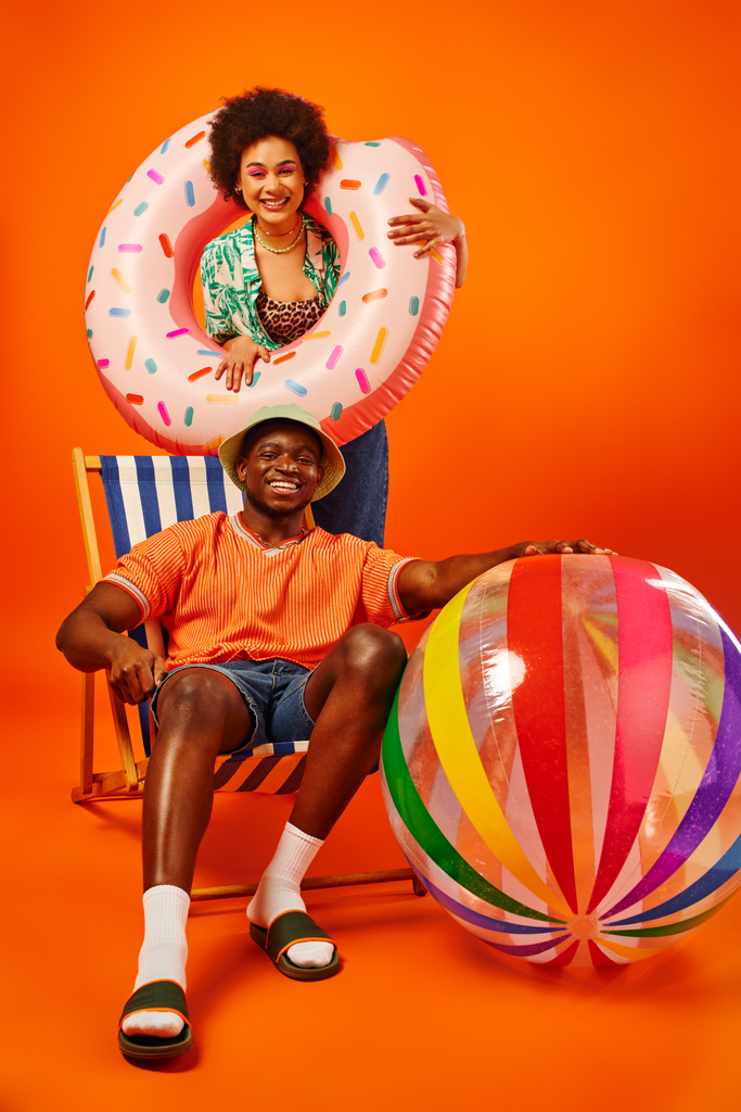 Full length of positive african american woman holding swim ring while standing near best friend in panama hat and summer outfit sitting on deck chair on orange background, fashion-forward friends - Photo, Image