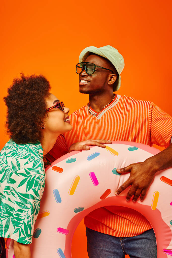 Potěšení mladí afroameričtí nejlepší přátelé ve slunečních brýlích a letních šatech drží plavecký prsten, tráví čas a stojí spolu izolované na oranžové, přátelé v módní ležérní oblečení - Fotografie, Obrázek