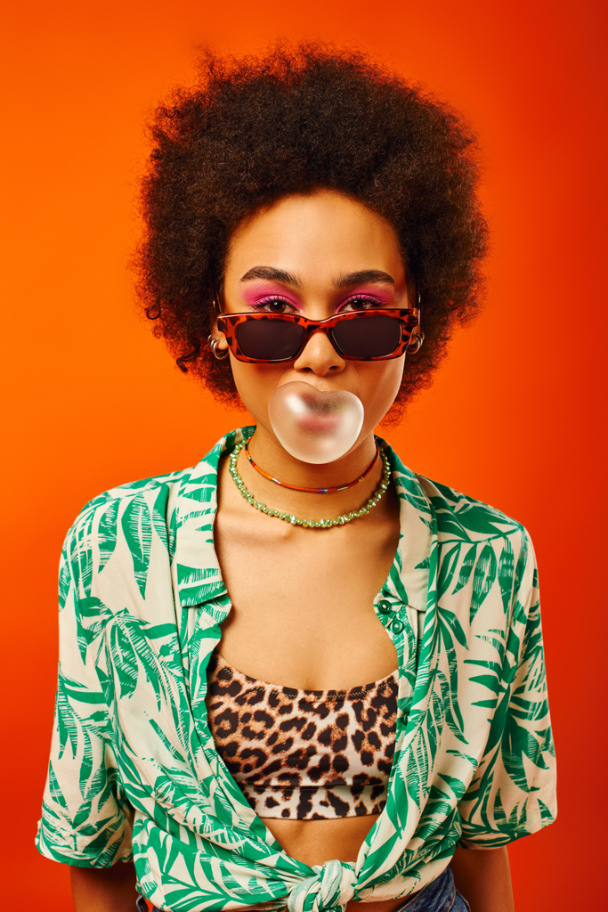 Portret młodej i modnej Afroamerykanki z odważnym makijażem w okularach przeciwsłonecznych i letnim stroju i dmuchającą gumą balonową wyizolowaną na czerwono, kobieta z poczuciem stylu, pewność siebie - Zdjęcie, obraz