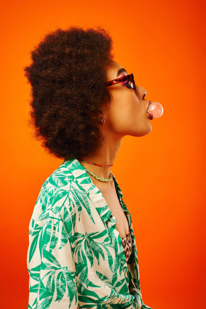 Vue latérale de jeune femme afro-américaine à la mode portant des lunettes de soleil en tenue d'été tout en soufflant de la gomme à bulles et se tenant isolée sur le rouge, femme avec un sens du style, confiance - Photo, image