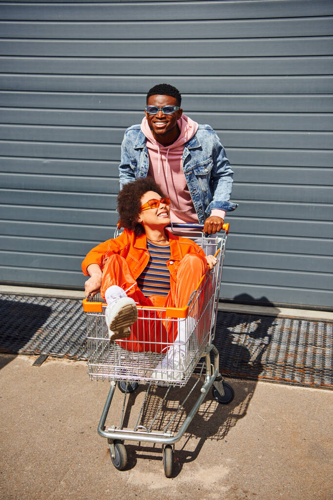 Hymyilevä nuori afrikkalainen amerikkalainen mies farkkutakissa ja aurinkolaseissa seisoo lähellä parasta ystävää ostoskärryissä ja rakennuksessa kaupunkien kadulla, ystävät hengailevat yhdessä, ystävyys - Valokuva, kuva