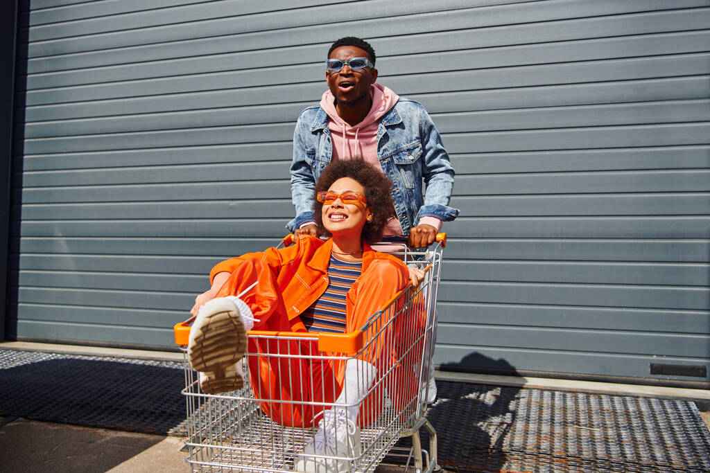 サングラスとデニムのジャケットを着た興奮した若いアフリカ系アメリカ人男性が笑顔の親友の近くに立ってショッピングカートに座って、都会の通りに建物、友人が一緒にぶらついて - 写真・画像