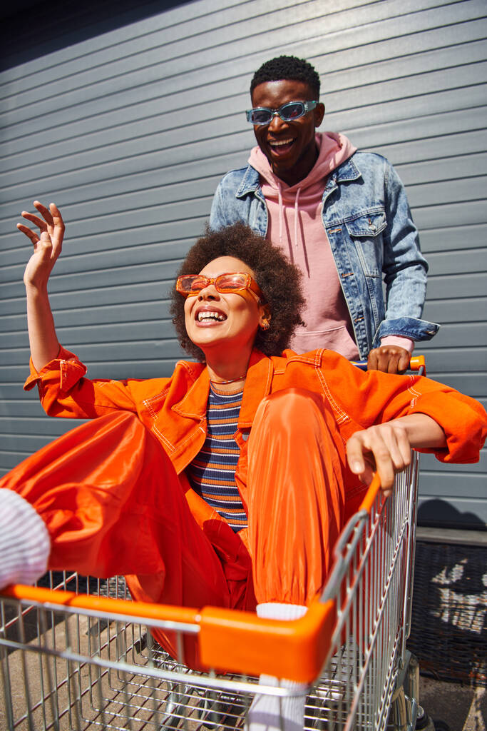 Veselý mladý africký Američan žena v jasném oblečení a sluneční brýle sedí v nákupním košíku a baví se s nejlepším přítelem v blízkosti budovy na městské ulici, přátelé visí spolu - Fotografie, Obrázek