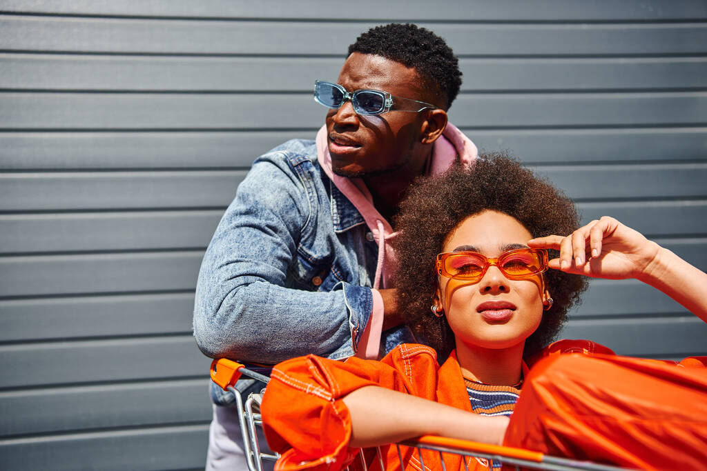 Itsevarma ja trendikäs nuori afrikkalainen amerikkalainen parhaita ystäviä aurinkolasit ja kirkkaat vaatteet poseeraa ostoskorin lähellä rakennuksen taustalla kaupunkien kadulla, ystäviä tyylikäs tunnelma - Valokuva, kuva