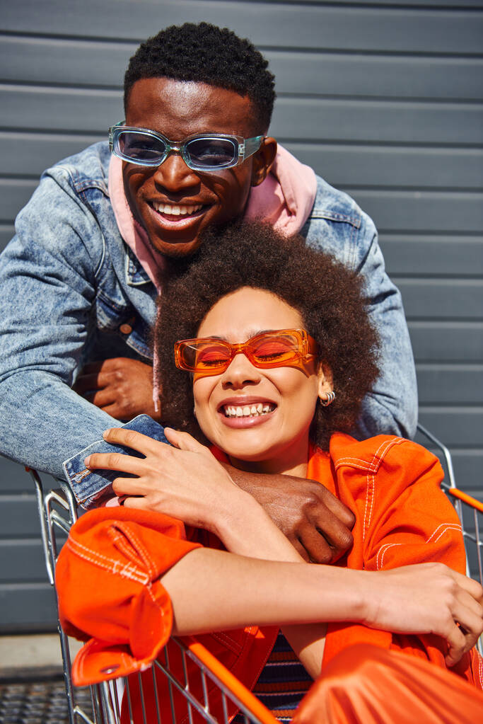 Ritratto di giovane uomo afroamericano allegro in occhiali da sole e giacca di jeans guardando la macchina fotografica mentre abbraccia il migliore amico nel carrello della spesa vicino all'edificio sulla strada urbana, amici con un'atmosfera elegante - Foto, immagini