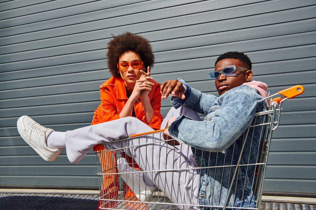 Kendine güvenen ve modaya uyan Afro-Amerikalı güneş gözlüklü ve parlak elbiseli en iyi arkadaşlar şehir caddesindeki binanın yakınında alışveriş arabasıyla poz veriyorlar. - Fotoğraf, Görsel