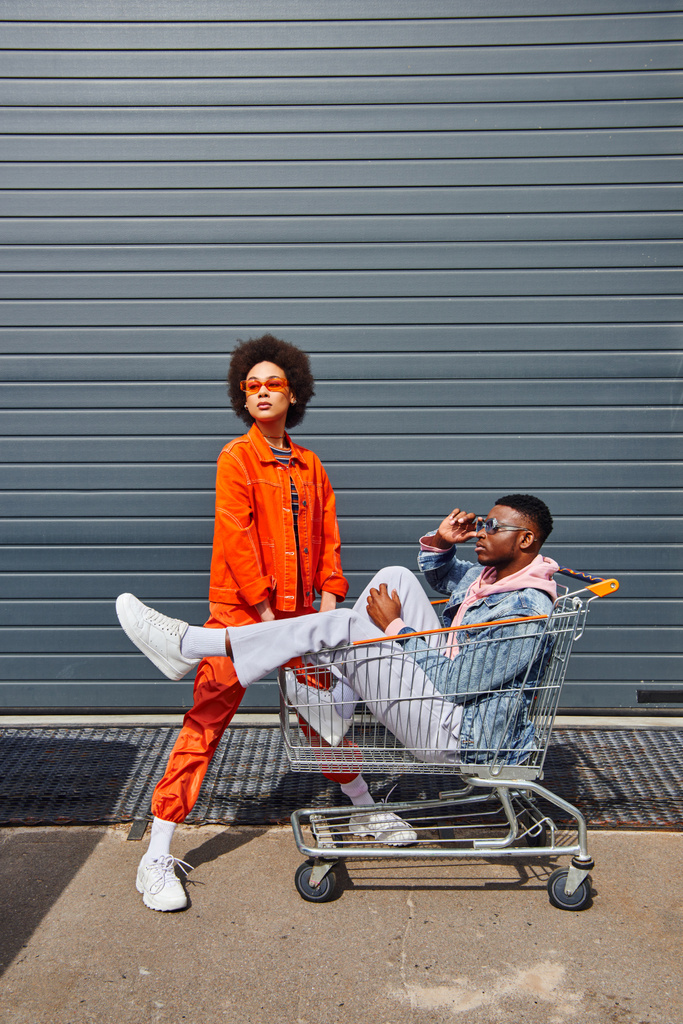 Plná délka módní mladý africký Američan muž nosí sluneční brýle, zatímco sedí v nákupním košíku poblíž nejlepší přítel v jasném oblečení a stavební venku, přátelé se stylovou atmosférou - Fotografie, Obrázek