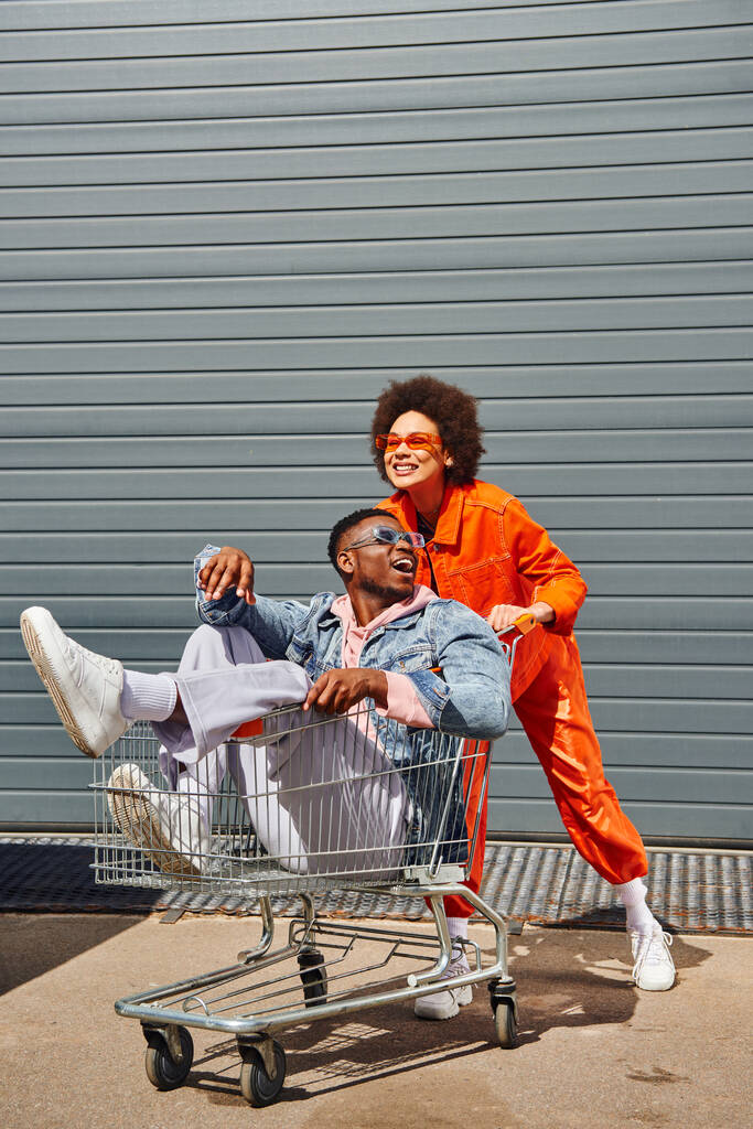 Jovens e animados afro-americanos melhores amigos em óculos de sol e roupas brilhantes se divertindo com carrinho de compras e passar o tempo perto de construção na rua urbana, amigos com vibe elegante - Foto, Imagem