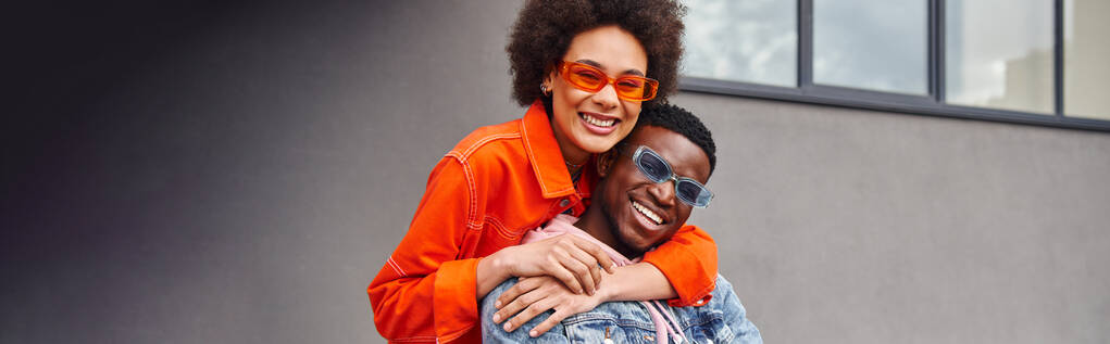 Pozytywna młoda Afroamerykanka w okularach przeciwsłonecznych i jasnym stroju obejmująca najlepszą przyjaciółkę i patrząca w kamerę, stojąca w pobliżu budynku na ulicy miejskiej, stylowe przyjaciółki w mieście, sztandar  - Zdjęcie, obraz