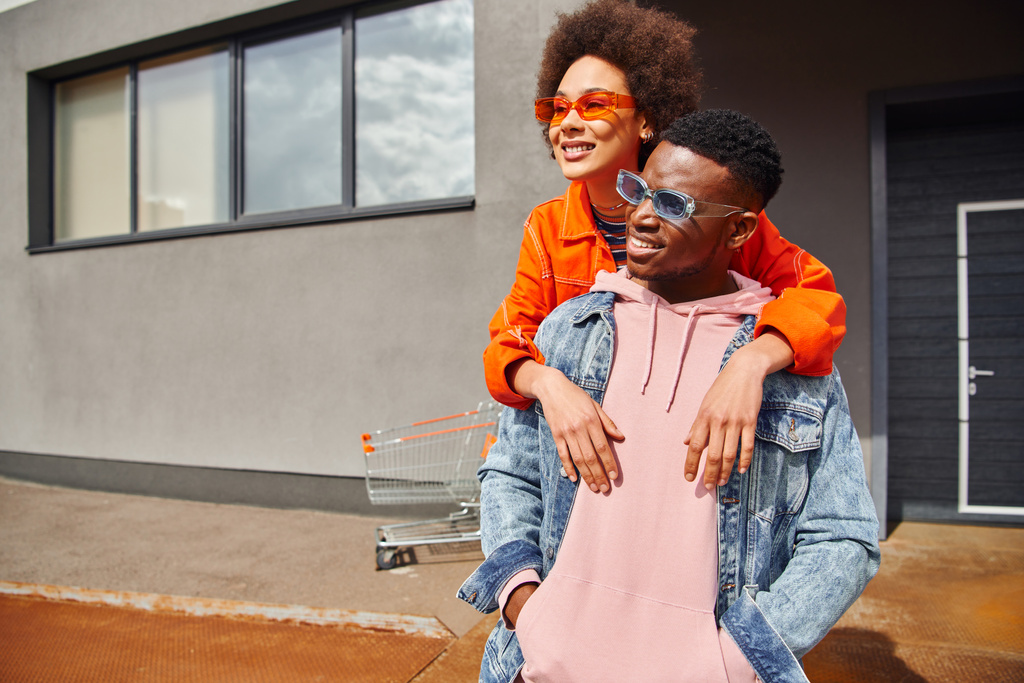 Glimlachende jonge Afro-Amerikaanse vrouw in zonnebril en heldere kleren omarmen modieuze beste vriend in denim jas, terwijl het doorbrengen van tijd op stedelijke straat, stijlvolle vrienden in de stad concept - Foto, afbeelding