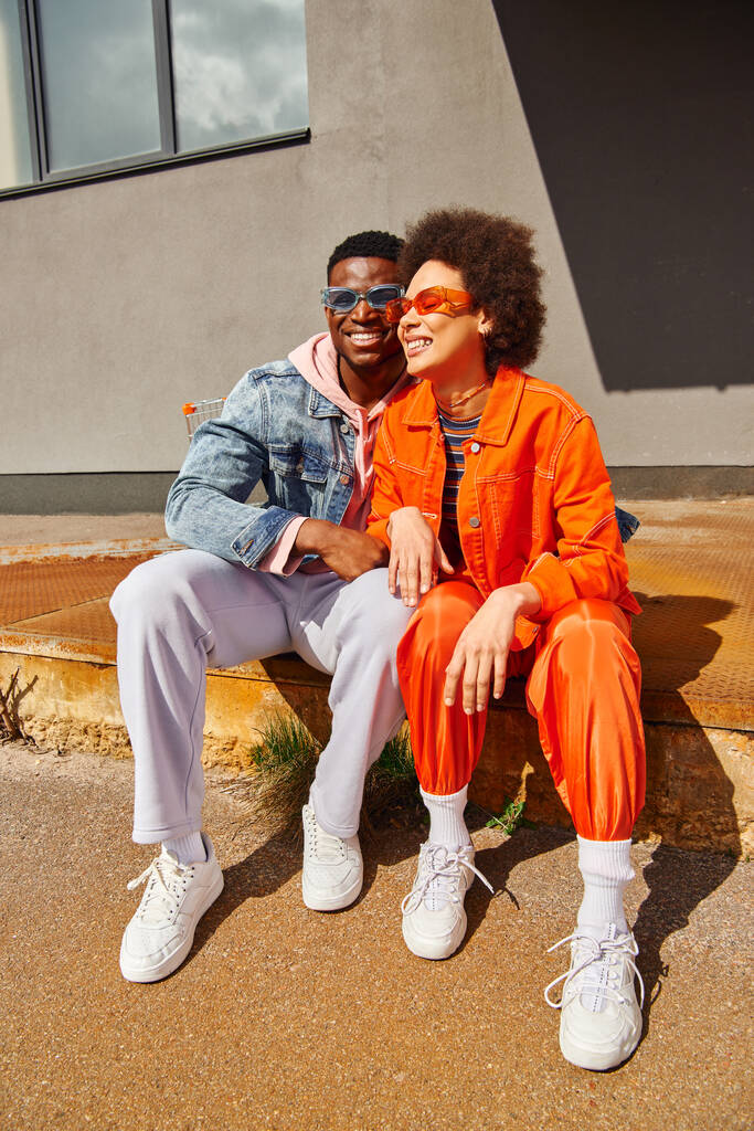 Sonriente joven afroamericano hombre en gafas de sol abrazando a la moda mejor amigo en traje brillante mientras está sentado en escaleras oxidadas cerca del edificio en la calle urbana, amigos con estilo en la ciudad - Foto, Imagen