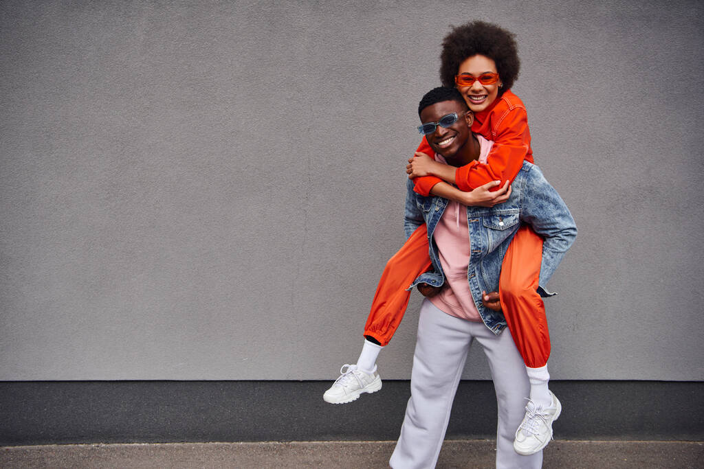 Smiling young african american woman in sunglasses and bright outfit piggybacking on stylish best friend and looking at camera near building on urban street, trendy friends in urban settings - Photo, Image