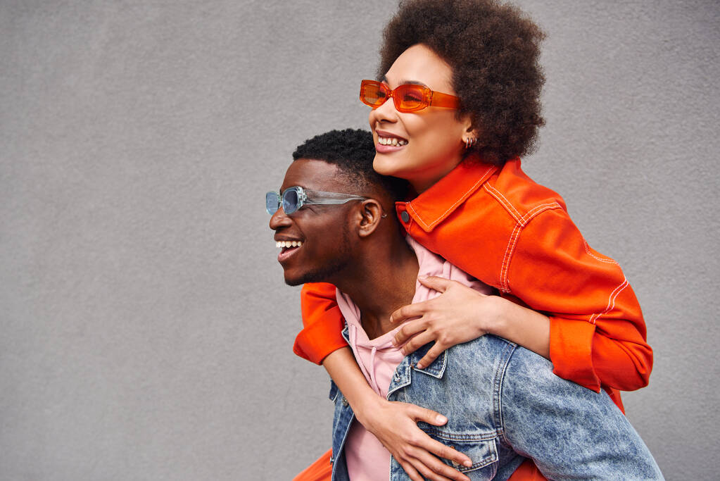 Young and stylish african american woman in sunglasses piggybacking on best friend in denim jacket and having fun near building on urban street, trendy friends in urban settings - Photo, Image