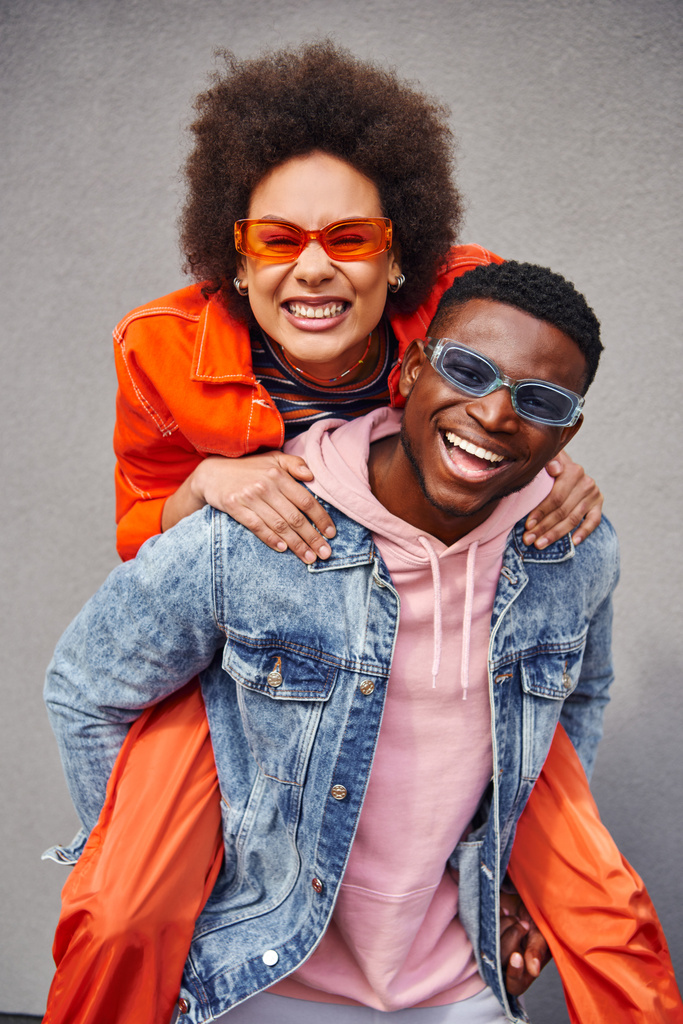 Retrato de una joven afro-americana positiva y con estilo en gafas de sol apoyándose en el mejor amigo en chaqueta de mezclilla y mirando a la cámara cerca del edificio al aire libre, amigos de moda en entornos urbanos - Foto, imagen