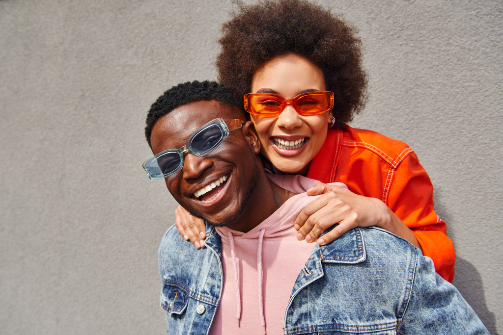 Ritratto di giovane donna afroamericana allegra con capelli naturali che abbraccia il migliore amico in occhiali da sole alla moda e giacca di jeans mentre guarda la fotocamera vicino all'edificio, amici alla moda in ambienti urbani - Foto, immagini