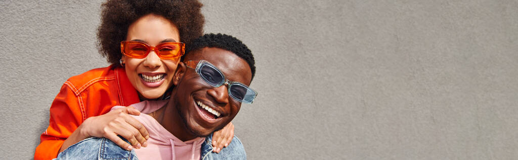 Fröhliche afrikanisch-amerikanische Frau in moderner Sonnenbrille und hellem Outfit umarmt beste Freundin und blickt in die Kamera, während sie in der Nähe eines Gebäudes auf der städtischen Straße steht, Freunde mit trendiger Ästhetik, Banner - Foto, Bild