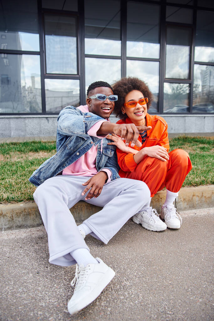 Usmívající se a trendy mladý africký Američan ve slunečních brýlích ukazuje prstem, zatímco sedí na hranici vedle nejlepšího přítele ve stylovém oblečení na městské ulici, přátelé s trendy estetiky - Fotografie, Obrázek