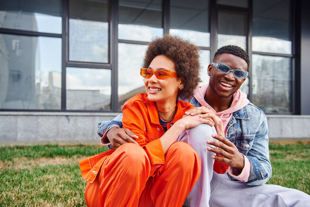 陽気で若いスタイリッシュなアフリカ系アメリカ人の男性がサングラスをかけて明るい服で親友を抱き、都会の通りのぼやけた建物の近くに草の上に座って、流行の美学を持つ友人 - 写真・画像