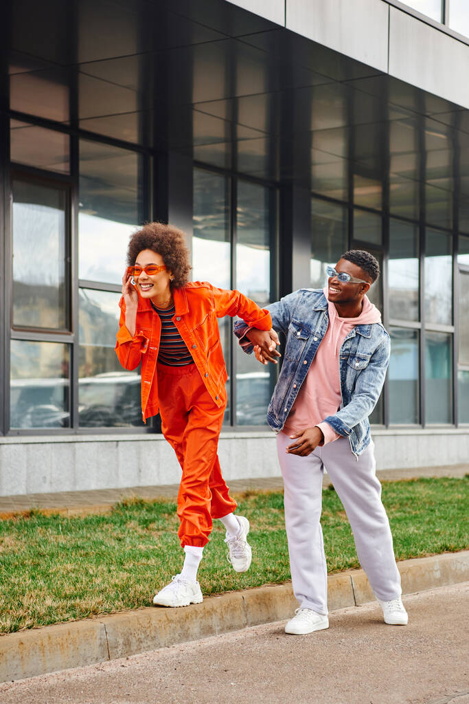 Πλήρες μήκος των χαρούμενα και σύγχρονα αφροαμερικανός γυναίκα σε γυαλιά ηλίου και φωτεινό ντύσιμο με τα πόδια στα σύνορα και κρατώντας το χέρι του καλύτερου φίλου σε αστικό δρόμο, κομψό φίλους απολαμβάνοντας την εταιρεία - Φωτογραφία, εικόνα
