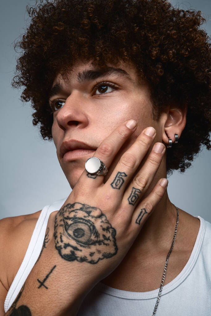 Ethnic male bandit with Afro hair touching cheek with tattooed hand looking away on gray background - 写真・画像