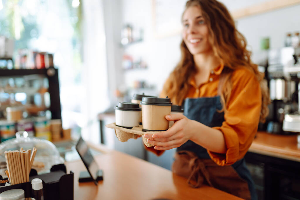 A legjobb kávé neked. Vidám nő kötényben a bárpultnál kávéfőzőpoharakat tart egy kávézóban. Elvitt élelmiszer fogalma. - Fotó, kép