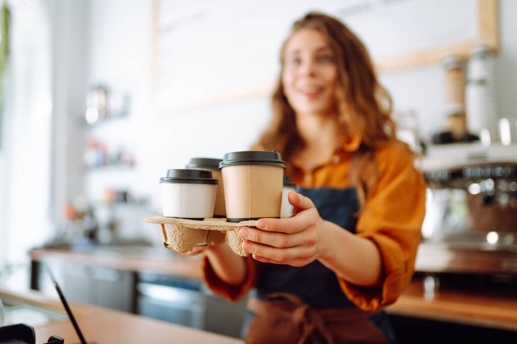 Der beste Kaffee für Sie. Gut gelaunte Frau in Schürze am Tresen hält in einem Café Kaffeegläser in der Hand. Konzept Essen zum Mitnehmen. - Foto, Bild