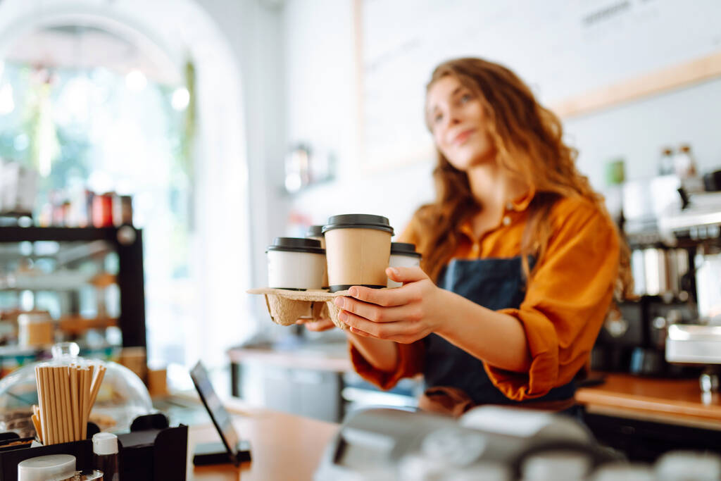 El mejor café para ti. Mujer alegre en un delantal en el mostrador del bar tiene vasos de café en un café. Concepto de comida para llevar. - Foto, imagen
