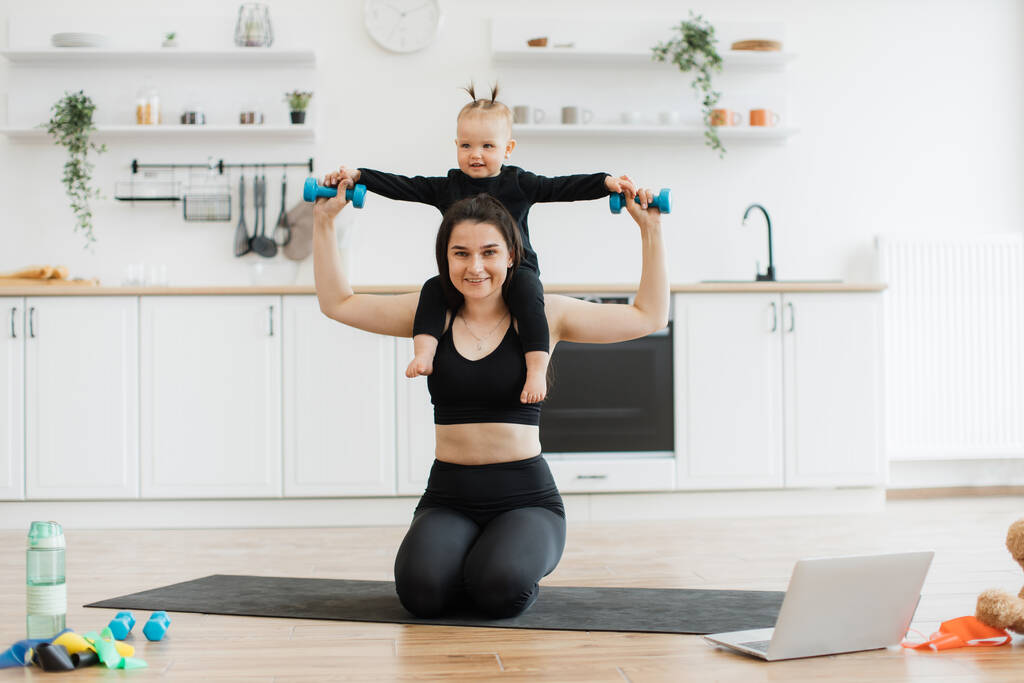 Joyful woman in yoga clothing carrying baby girl with dumbbells on shoulders while sitting on knees indoors. Sporty mother and daughter turning daily workout into amusing entertainment at home. - Photo, Image