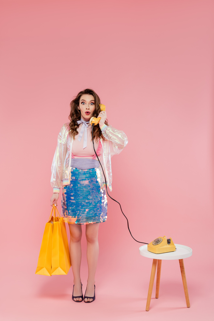 έκπληκτη νεαρή γυναίκα που μεταφέρει τσάντες για ψώνια και μιλάει σε ρετρό τηλέφωνο, στέκεται σε ροζ φόντο, εννοιολογική φωτογραφία, τηλεφωνική κλήση, vintage τηλέφωνο, έννοια νοικοκυρά  - Φωτογραφία, εικόνα