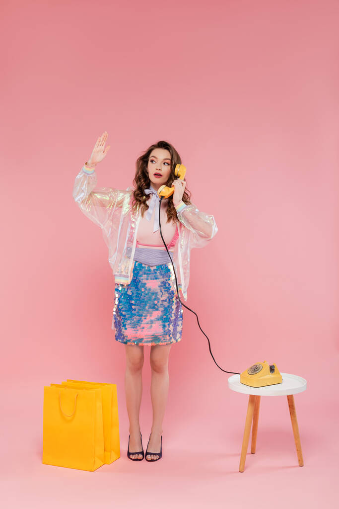 hermosa mujer que tiene llamada telefónica, posando cerca de bolsas de compras, de pie como una muñeca y sosteniendo el auricular retro sobre fondo rosa, fotografía conceptual, pose de muñeca, ama de casa mirando las uñas  - Foto, Imagen